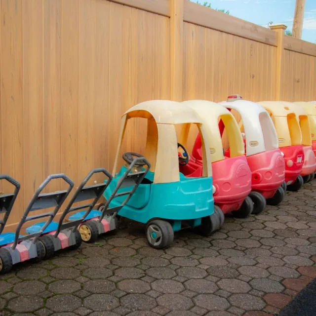 Toy cars and toy lawn mowers lined up on a sidewalk next to a tall wooden fence.