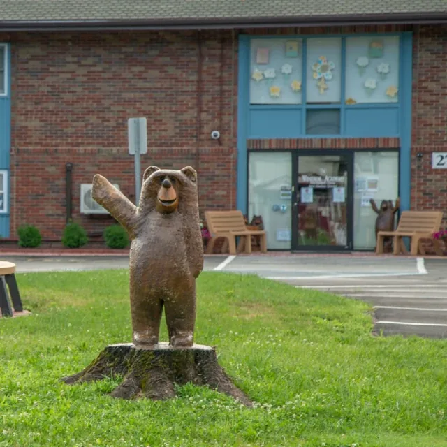 A wooden bear statue on a tree stump waves near a building with decorated windows.