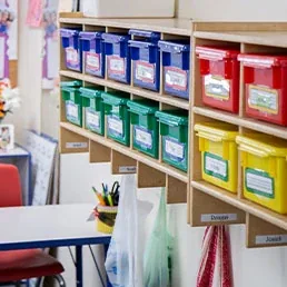 Colorful plastic bins on shelves in a classroom, with name labels and supplies below.