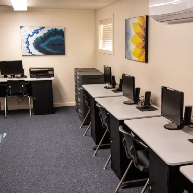 Small computer lab with desks, computers, chairs, wall art, and a printer in the corner.