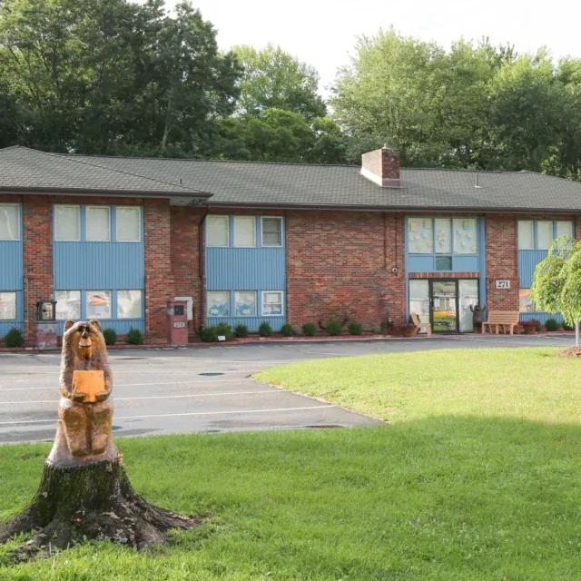 Two-story brick building with blue panels, trees, and a carved bear statue on a tree stump.