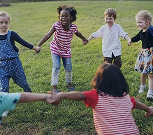 Children holding hands in a circle and playing on grass, smiling and enjoying outdoor time.
