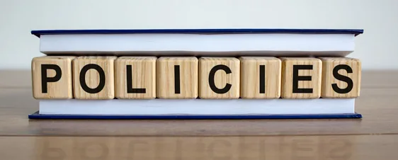 Wooden blocks spelling POLICIES are stacked on top of a closed book on a wooden table.