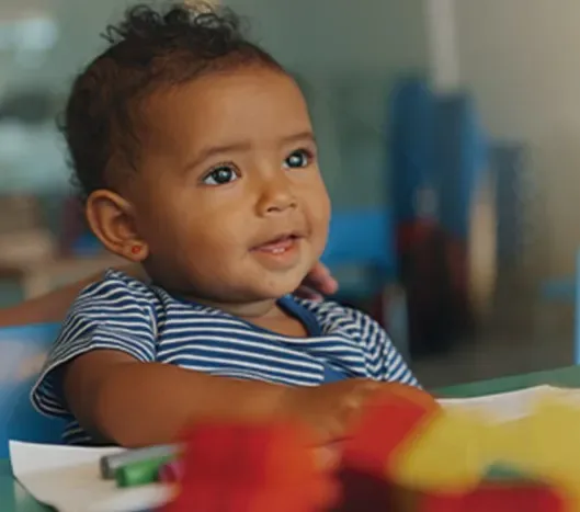 Smiling toddler in a striped shirt sitting at a table with colorful toys and papers.