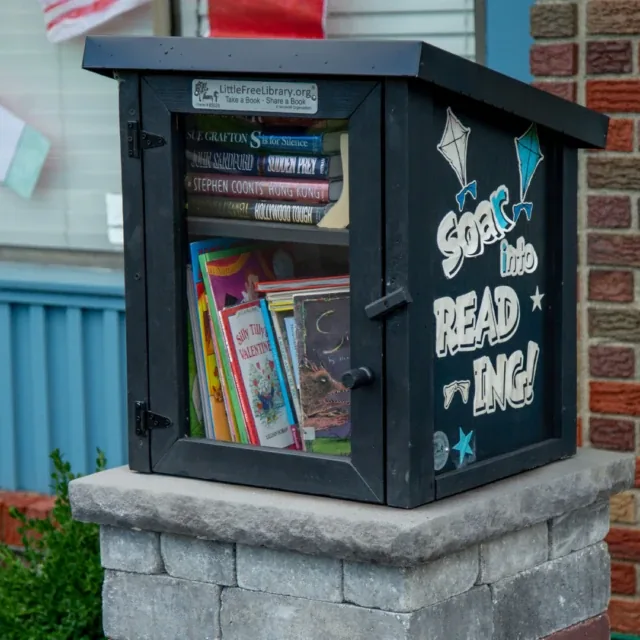 A little free library box filled with books sits on a stone pedestal outside a building.