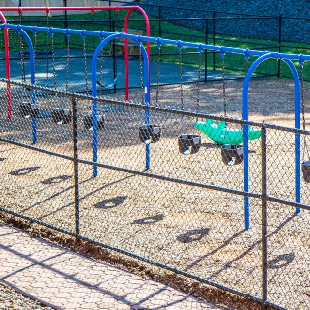 Empty swings behind a chain-link fence at a playground on a sunny day.