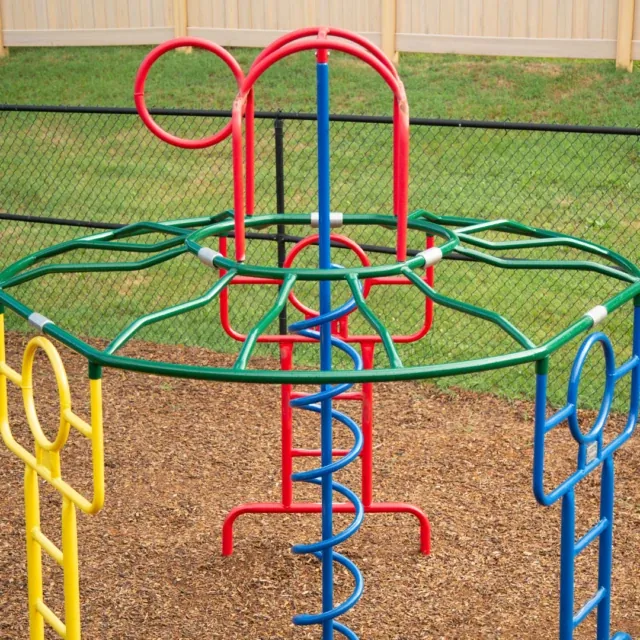 Colorful metal climbing structure at a playground with mulch ground and a fenced background.