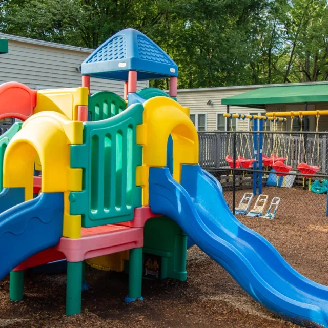 Colorful playground with slides and swings outdoors near a building, surrounded by trees.