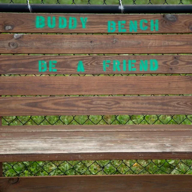 A wooden bench with green text that reads, Buddy Bench Be a Friend, in a fenced area.