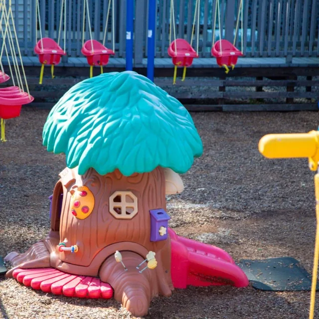 Plastic playhouse shaped like a tree with a green roof, surrounded by red swings at a playground.