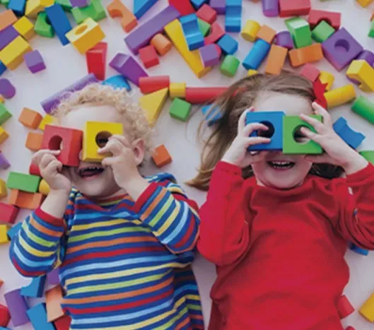 Two children playing with colorful blocks and holding some up to their eyes, smiling and laughing.