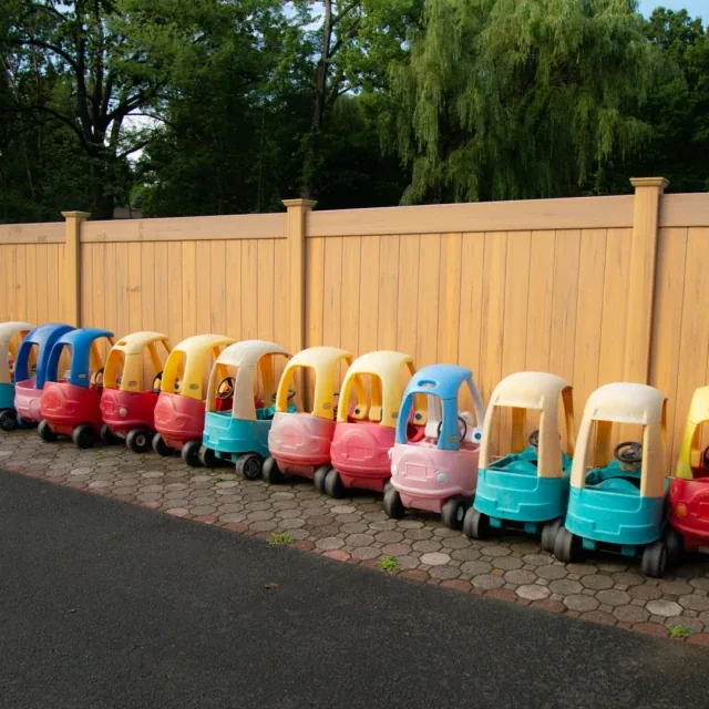 A row of colorful toy cars is lined up along a wooden fence on a paved surface.