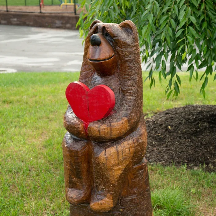 Wooden bear statue sitting and holding a red heart, positioned outdoors on grass near a tree.