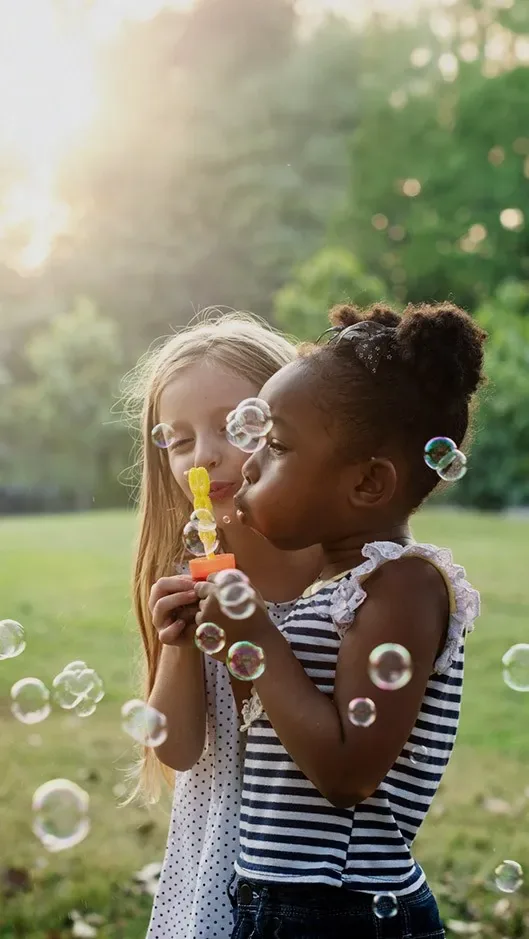 Two young girls blow bubbles together outside on a sunny day, surrounded by green trees.