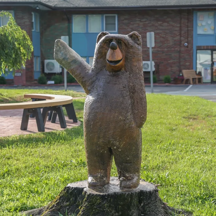 Wooden bear statue with one paw raised, standing on a tree stump in a grassy area.