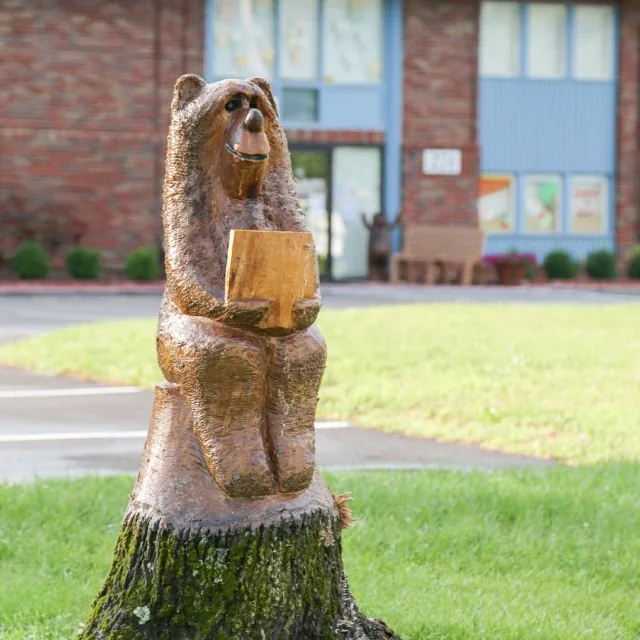 Wooden bear statue carved from a tree stump, holding a book, on grass near a brick building.