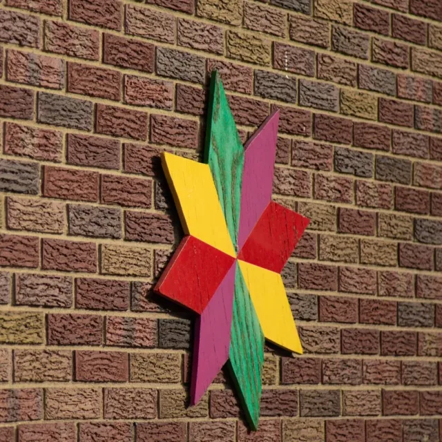 A colorful wooden star with eight points hangs on a textured brick wall.