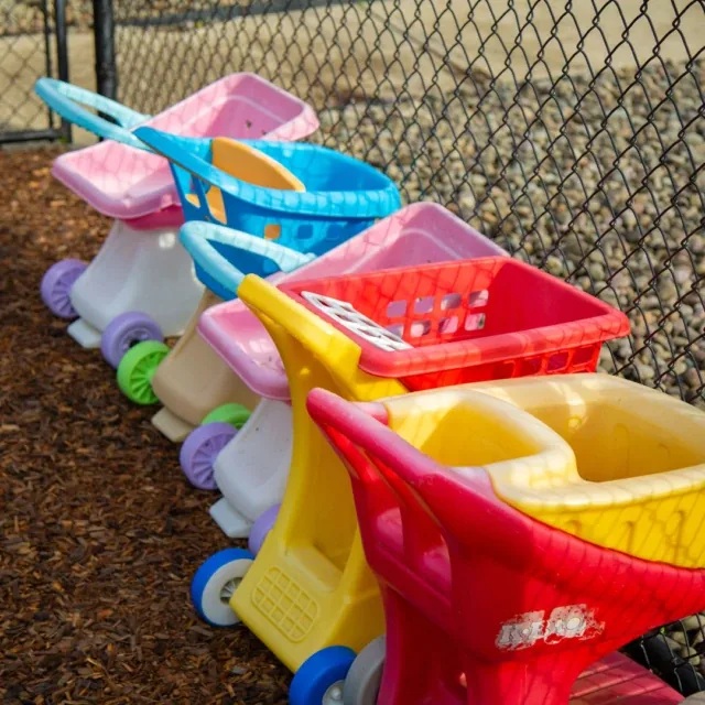 Five colorful toy shopping carts are lined up along a chain-link fence on a playground.