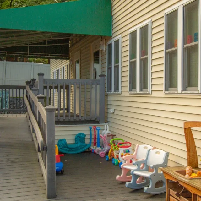 Childrens plastic toys and chairs are grouped on a porch beside a beige house with large windows.