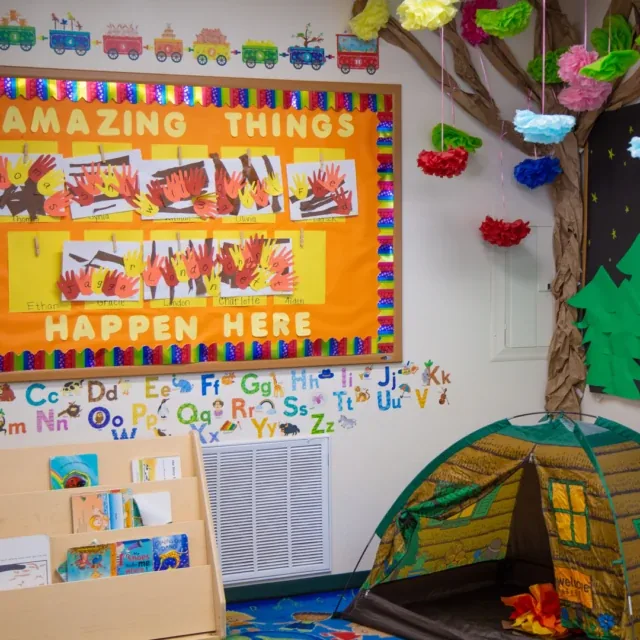 Colorful classroom with a bulletin board, book rack, alphabet wall, and a play tent under a tree.