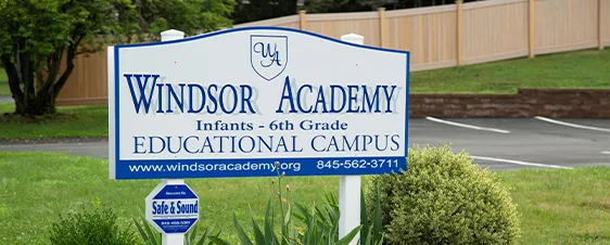A sign for Windsor Academy, infants to 6th grade educational campus, near a road and greenery.