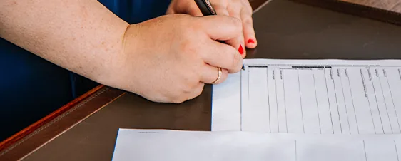 A person with red nail polish fills out a form on a desk with a pen.