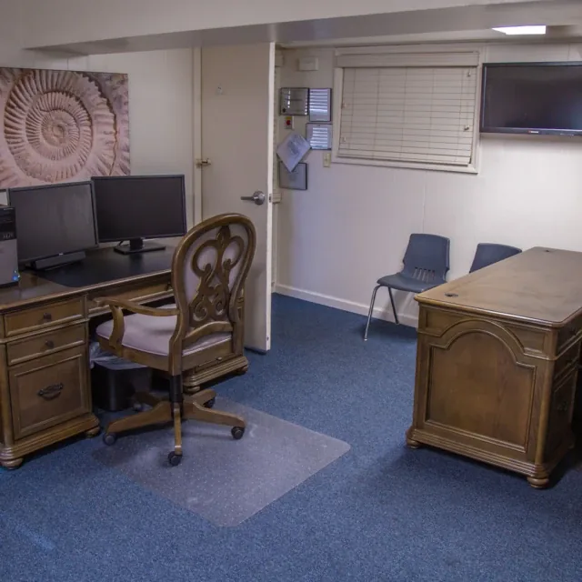 Office with two wooden desks, computer monitors, chairs, wall art, and a mounted TV.